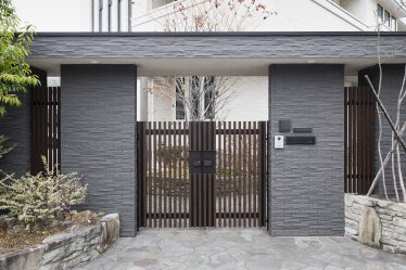 Nowoczesne wejście do posesji z dwuskrzydłową, drewnianą furtką w stylu japońskim, otoczoną szarym murem z klinkieru i eleganckim ogrodzeniem – przestrzeń idealna na napęd do bramy dwuskrzydłowej FAAC.