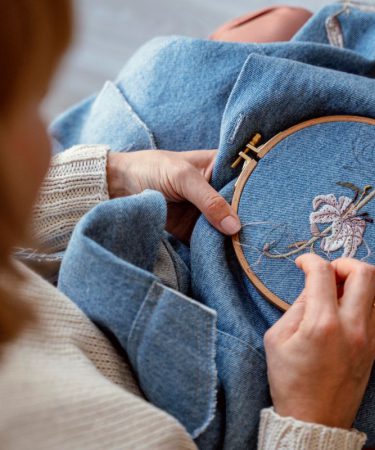 Dłonie kobiety wyszywającej jasny kwiat na niebieskiej tkaninie za pomocą tamborka i igły. Zdjęcie pokazuje, jak powstają detale na koszulkach damskich z haftem.