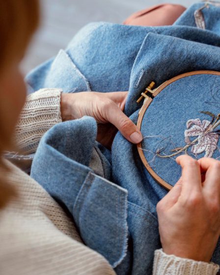 Dłonie kobiety wyszywającej jasny kwiat na niebieskiej tkaninie za pomocą tamborka i igły. Zdjęcie pokazuje, jak powstają detale na koszulkach damskich z haftem.
