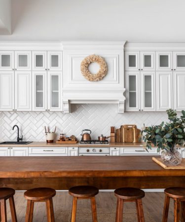 Projekt kuchni z jadalnią, w którym biała zabudowa z przeszklonymi szafkami łączy się z dużą, drewnianą wyspą pełniącą funkcję stołu. Na ścianie nad kuchenką widoczne są białe płytki w jodełkę, a w centralnej części wyspy stoi wazon z zielonymi gałązkami eukaliptusa.