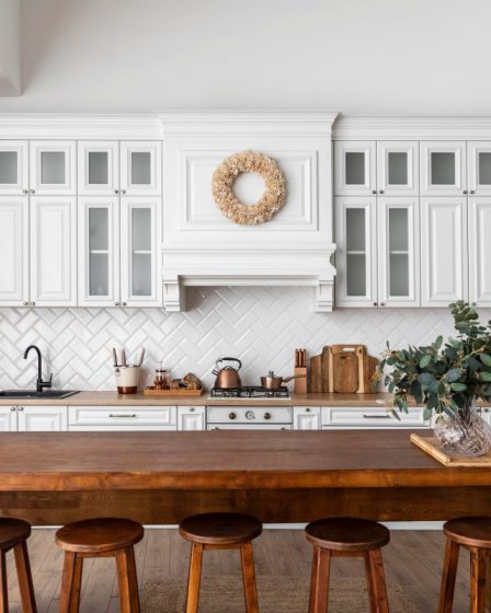 Projekt kuchni z jadalnią, w którym biała zabudowa z przeszklonymi szafkami łączy się z dużą, drewnianą wyspą pełniącą funkcję stołu. Na ścianie nad kuchenką widoczne są białe płytki w jodełkę, a w centralnej części wyspy stoi wazon z zielonymi gałązkami eukaliptusa.