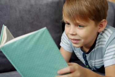 Na zdjęciu widać chłopca leżącego na kanapie, który z zaciekawieniem czyta książkę o zielonej okładce.