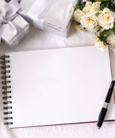 Otwarta biała księga gości na spiralach, leżąca na koronkowym obrusie obok eleganckiego pióra, srebrnych upominków i delikatnych kremowych kwiatów.