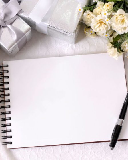 Otwarta biała księga gości na spiralach, leżąca na koronkowym obrusie obok eleganckiego pióra, srebrnych upominków i delikatnych kremowych kwiatów.