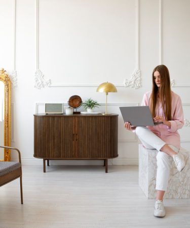 Młoda kobieta pracuje na laptopie w jasnym pokoju o klasycznym wystroju z ozdobną sztukaterią. Aranżacja stworzona przez studio projektowania wnętrz obejmuje stylową komodę, złote dodatki oraz lustro w ozdobnej ramie.