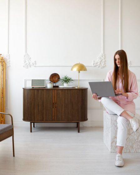 Młoda kobieta pracuje na laptopie w jasnym pokoju o klasycznym wystroju z ozdobną sztukaterią. Aranżacja stworzona przez studio projektowania wnętrz obejmuje stylową komodę, złote dodatki oraz lustro w ozdobnej ramie.