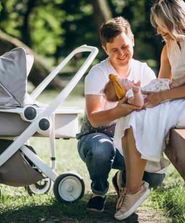 Uśmiechnięci rodzice siedzący na ławce i bawiący się z niemowlęciem obok nowoczesnej gondoli. Jasnoszare polskie wózki dziecięce na białym stelażu zaparkowane na trawie w słoneczny dzień.
