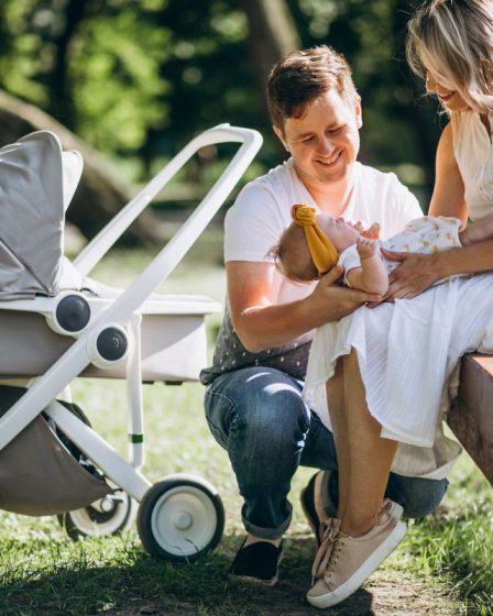 Uśmiechnięci rodzice siedzący na ławce i bawiący się z niemowlęciem obok nowoczesnej gondoli. Jasnoszare polskie wózki dziecięce na białym stelażu zaparkowane na trawie w słoneczny dzień.