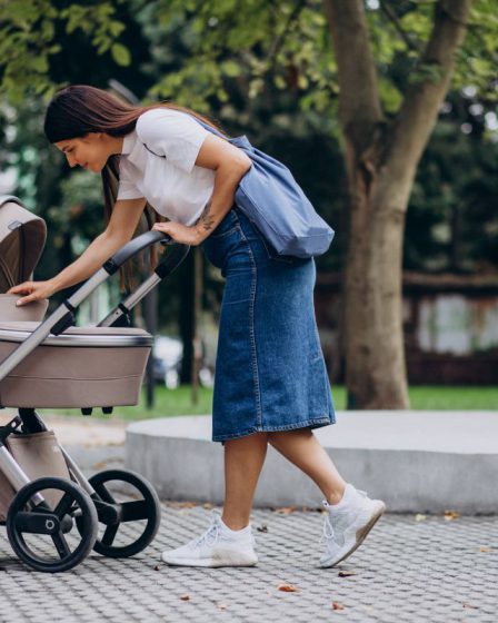 Kobieta spacerująca z niemowlęciem w parku. Klasyczne, beżowe wózki dla dzieci na brukowanej alejce w otoczeniu zieleni.