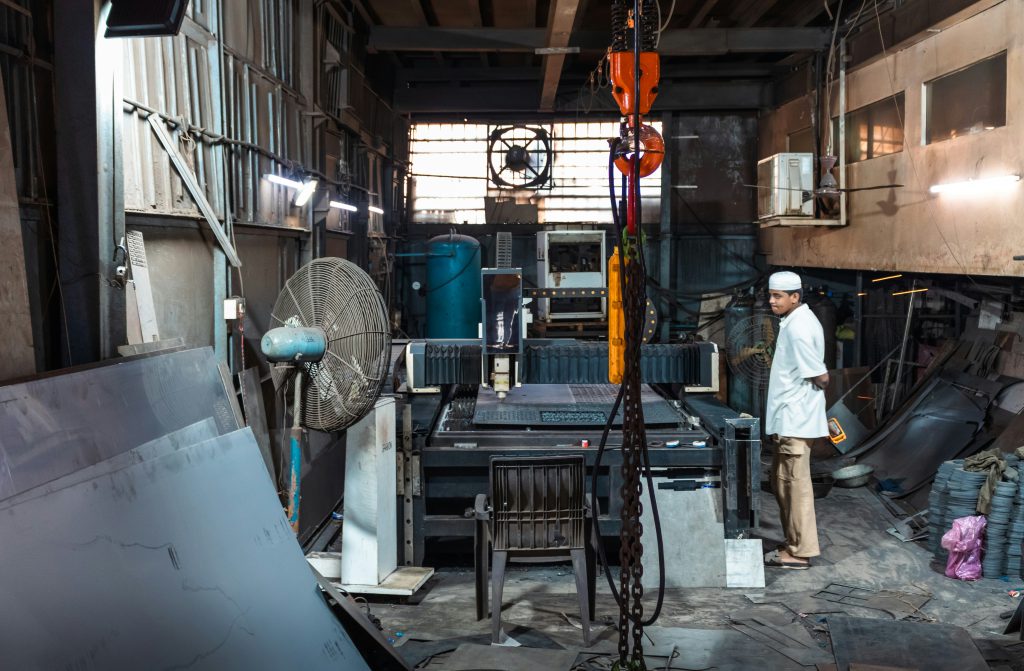 Na zdjęciu widać wnętrze warsztatu lub hali przemysłowej z dużą maszyną do obróbki metalu ustawioną w centralnej części pomieszczenia.